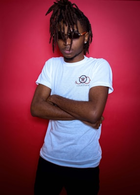 A person with long dreadlocks and sunglasses is standing against a vibrant red background. They are wearing a white T-shirt with a design on the chest and have their arms crossed.