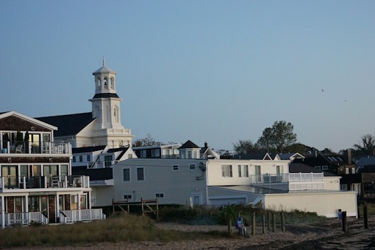A coastal scene with a white church in the background featuring a prominent tower, surrounded by various residential buildings with balconies. The foreground includes a sandy area with grass, as well as a person sitting on the sand. The sky is clear with a few birds flying.