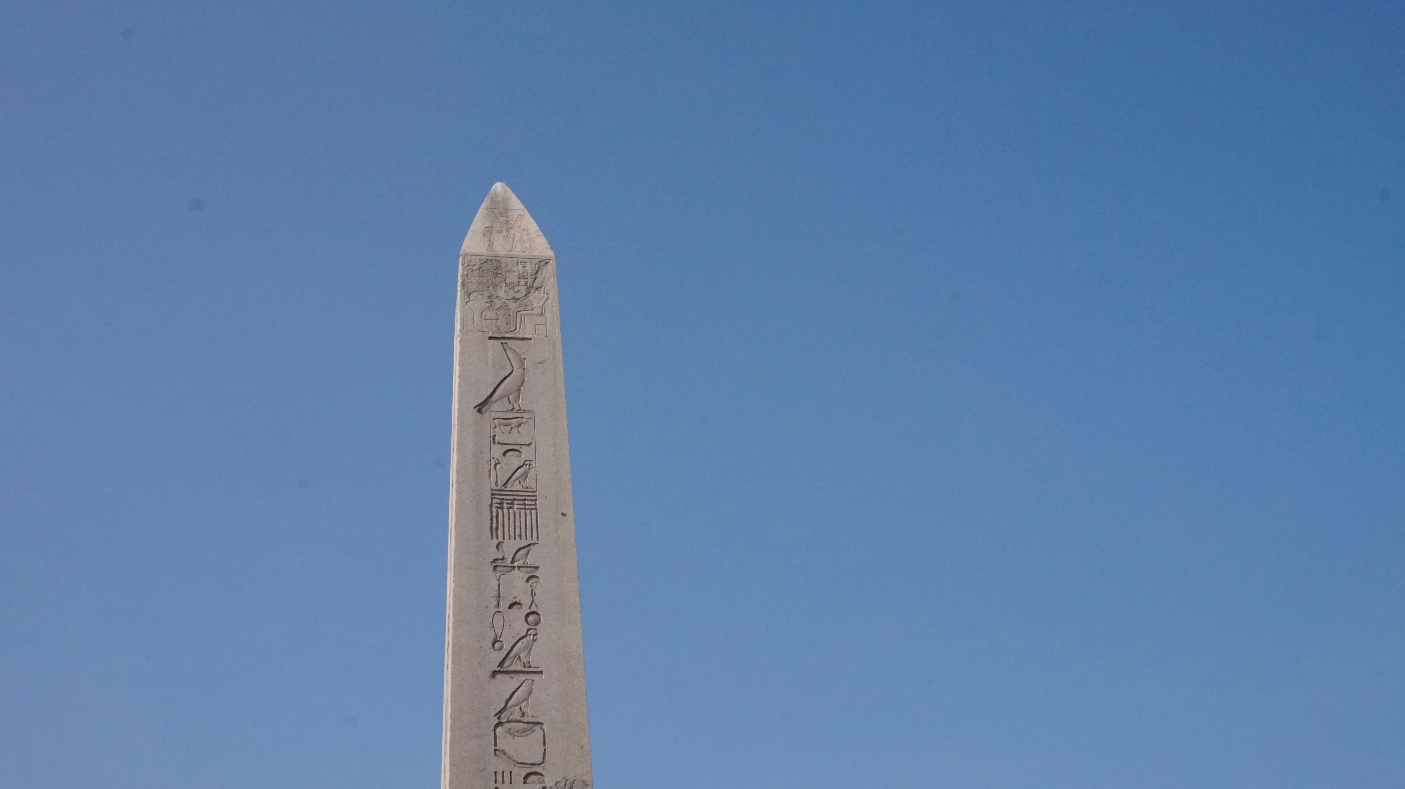 Ancient stone obelisk against blue sky, monumental scale, sunlit carved stone, historical grandeur