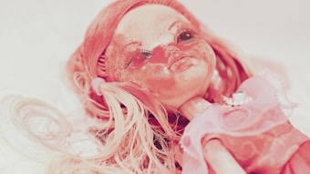 A close-up of a worn and slightly dirty doll with long, pinkish-blonde hair. The doll has large, prominent eyes with long lashes and a noticeable expression. It is dressed in a pink outfit and appears to have a vintage or slightly eerie aesthetic.