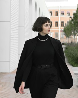 A person with short dark hair is walking through an outdoor urban area. They are wearing a black outfit consisting of a turtleneck, blazer, and high-waisted pants. The background features a modern building with white arches and a brick pathway. Some greenery is visible along the path.