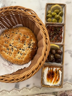A large round flatbread sprinkled with sesame and black seeds is placed in a woven basket. Adjacent to it, a rectangular set of ceramic dishes contains green olives garnished with lemon slices, mixed olives, a spicy salsa-like mixture, and a creamy dip drizzled with olive oil and garnished with spices.
