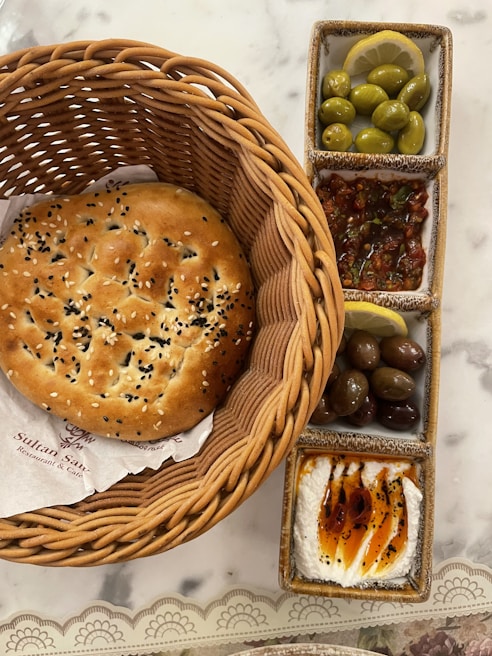 A large round flatbread sprinkled with sesame and black seeds is placed in a woven basket. Adjacent to it, a rectangular set of ceramic dishes contains green olives garnished with lemon slices, mixed olives, a spicy salsa-like mixture, and a creamy dip drizzled with olive oil and garnished with spices.
