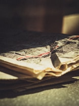 A close-up view of a crumpled newspaper featuring printed text. The paper is aged or weathered, with a yellowish tint. Shadows and light create a dramatic effect across the surface.