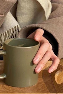 A close-up of a hand-touching a smooth, earthy-toned ceramic mug against a cozy wooden background.