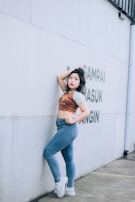 A person stands confidently against a light-colored wall with some text. They are wearing jeans, a cropped shirt, and white sneakers, with one arm resting on the wall and the other hand on the hip.