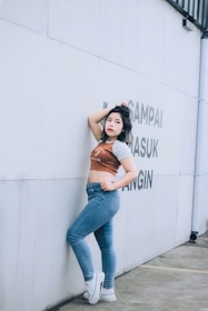 A person stands confidently against a light-colored wall with some text. They are wearing jeans, a cropped shirt, and white sneakers, with one arm resting on the wall and the other hand on the hip.