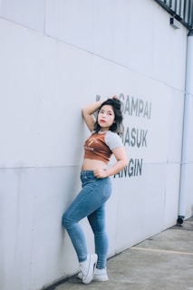 A person stands confidently against a light-colored wall with some text. They are wearing jeans, a cropped shirt, and white sneakers, with one arm resting on the wall and the other hand on the hip.