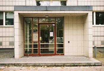 An entrance to a building with a glass door framed in red and a number 20 above it. The facade is covered in beige tiles, with a small awning above the door. There are scattered leaves on the ground in front of the entrance. A few windows line the upper portion of the building.