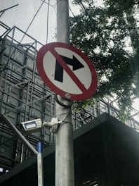 A no right turn traffic sign is mounted on a pole, accompanied by a surveillance camera. The background consists of a building with a metal structure and some tree branches partially visible.