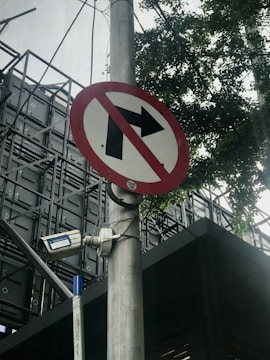 A no right turn traffic sign is mounted on a pole, accompanied by a surveillance camera. The background consists of a building with a metal structure and some tree branches partially visible.