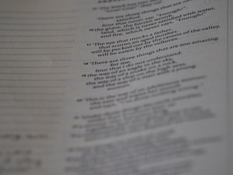 A page from a book with printed text, featuring numbered verses that appear poetic. The text is aligned along the center, and the image is slightly out of focus, particularly towards the bottom. The paper has a faint texture, with visible lines to the left side.