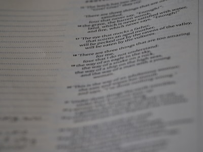 A page from a book with printed text, featuring numbered verses that appear poetic. The text is aligned along the center, and the image is slightly out of focus, particularly towards the bottom. The paper has a faint texture, with visible lines to the left side.