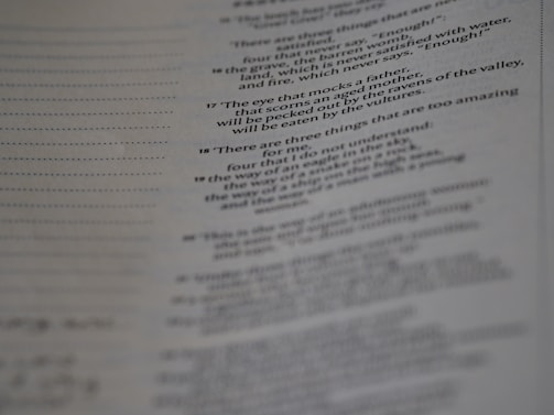 A page from a book with printed text, featuring numbered verses that appear poetic. The text is aligned along the center, and the image is slightly out of focus, particularly towards the bottom. The paper has a faint texture, with visible lines to the left side.