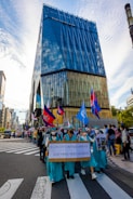 A vibrant city street parade featuring a group of people dressed in matching teal outfits, resembling the Statue of Liberty. They hold a sign indicating the 31st Paris Peace Accords. Various flags are being waved, including some with Cambodian insignia. The modern high-rise building named 'Tokyu Plaza' is prominently visible, reflecting the clear blue sky.
