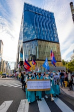 A vibrant city street parade featuring a group of people dressed in matching teal outfits, resembling the Statue of Liberty. They hold a sign indicating the 31st Paris Peace Accords. Various flags are being waved, including some with Cambodian insignia. The modern high-rise building named 'Tokyu Plaza' is prominently visible, reflecting the clear blue sky.