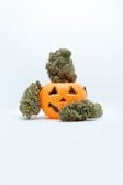 A small orange plastic jack-o'-lantern with a carved happy face is surrounded by several cannabis buds. One large bud is perched on top, while others are placed around the base.