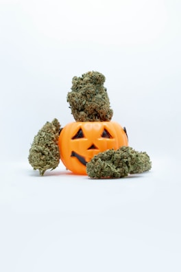 A small orange plastic jack-o'-lantern with a carved happy face is surrounded by several cannabis buds. One large bud is perched on top, while others are placed around the base.