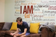 A young man sits on a brown couch with colorful pillows, appearing to contemplate or pray. Behind him is a wall covered in various religious words and phrases such as 'I AM', 'Prince of Peace', and 'Jesus'.