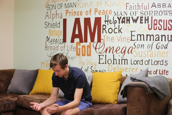 A young man sits on a brown couch with colorful pillows, appearing to contemplate or pray. Behind him is a wall covered in various religious words and phrases such as 'I AM', 'Prince of Peace', and 'Jesus'.