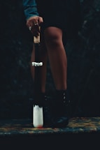A person stands with their legs slightly apart, holding a weapon resembling a baseball bat with a white tip and a handle wrapped in cord. The person's hand is adorned with rings and their outfit includes a denim jacket and black boots. The setting is dark and mysterious, heightening the dramatic effect.