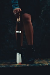 A person stands with their legs slightly apart, holding a weapon resembling a baseball bat with a white tip and a handle wrapped in cord. The person's hand is adorned with rings and their outfit includes a denim jacket and black boots. The setting is dark and mysterious, heightening the dramatic effect.