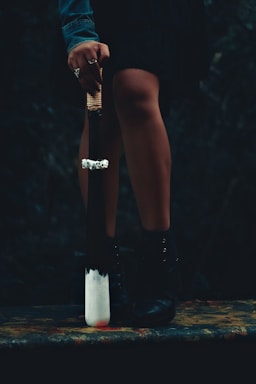 A person stands with their legs slightly apart, holding a weapon resembling a baseball bat with a white tip and a handle wrapped in cord. The person's hand is adorned with rings and their outfit includes a denim jacket and black boots. The setting is dark and mysterious, heightening the dramatic effect.