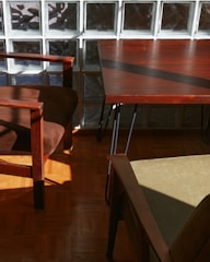 Sunlight streams onto a wooden floor, casting shadows from brown wooden furniture with soft cushions. A glass block wall forms the backdrop, enhancing the play of light and shadow.