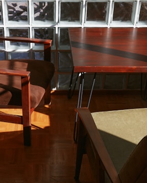 Sunlight streams onto a wooden floor, casting shadows from brown wooden furniture with soft cushions. A glass block wall forms the backdrop, enhancing the play of light and shadow.