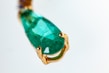 A close-up of a teardrop-shaped green gemstone set in a gold clasp. The gemstone has a smooth surface with visible natural inclusions, highlighting its authenticity. The gold setting has a rich, polished texture that complements the vibrant color of the stone.