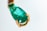 A close-up of a teardrop-shaped green gemstone set in a gold clasp. The gemstone has a smooth surface with visible natural inclusions, highlighting its authenticity. The gold setting has a rich, polished texture that complements the vibrant color of the stone.