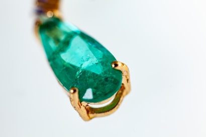 A close-up of a teardrop-shaped green gemstone set in a gold clasp. The gemstone has a smooth surface with visible natural inclusions, highlighting its authenticity. The gold setting has a rich, polished texture that complements the vibrant color of the stone.