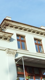 A classic architectural building facade with ornate details, including decorative cornices and pilasters. The exterior is a light color with large wooden-framed windows. A small balcony with a metal railing and some visible flowers can be seen.