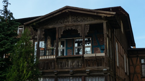 A traditional wooden house with intricate carvings on the balcony and a mix of exposed brickwork on the facade. Tall trees frame the scene, adding to the rustic and natural atmosphere.