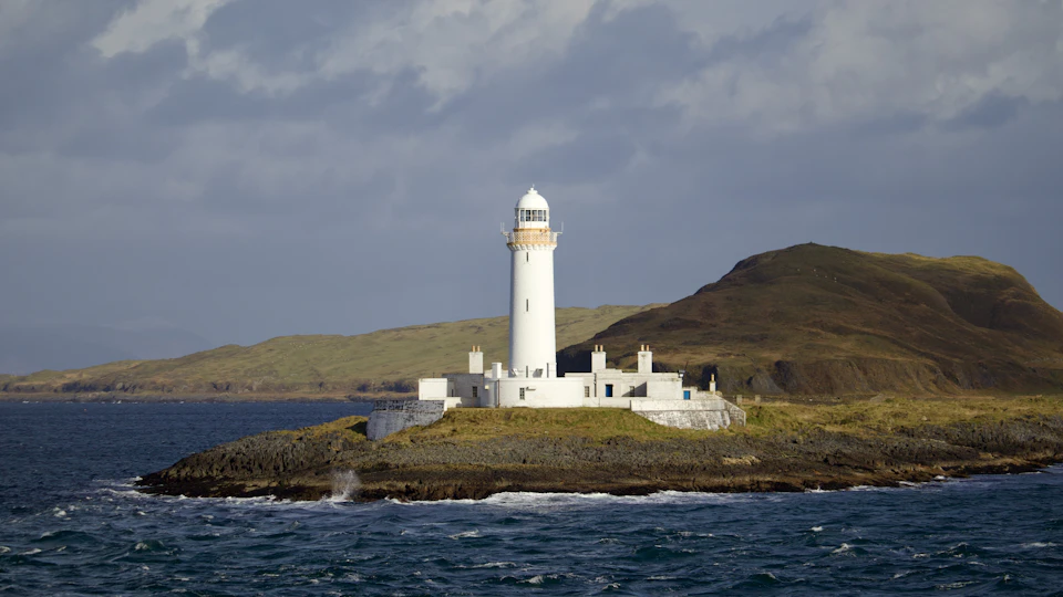 The image depicts a serene coastal scene featuring a white lighthouse standing tall on a rocky island, with its light glowing against an overcast sky. The lighthouse is surrounded by water and features multiple chimneys protruding from its sides. In the distance, there are hills visible in the background, adding depth to the landscape.