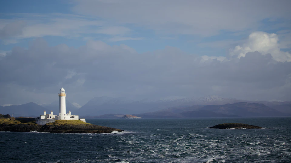 The image depicts a serene scene at sea, with a white lighthouse standing tall on a rocky island in the center. The lighthouse is surrounded by choppy waters and a cloudy sky, creating an atmosphere of tranquility. In the distance, mountains can be seen rising against the horizon, adding depth to the landscape.