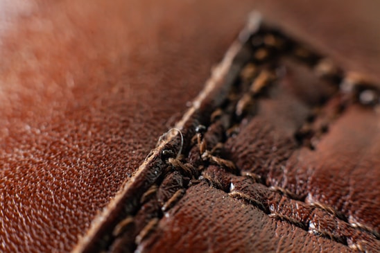 A close-up view of brown leather with detailed stitching running vertically. The texture of the leather is visible, showing its grainy surface and aged appearance.