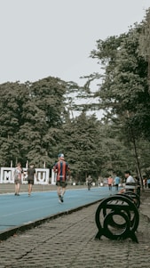 A vibrant outdoor scene featuring a running track surrounded by lush green trees. People are actively walking or jogging on the blue track, some dressed in sportswear. Benches and a decorative sculpture are visible alongside the track. The setting appears lively and recreational.