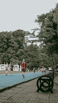 A vibrant outdoor scene featuring a running track surrounded by lush green trees. People are actively walking or jogging on the blue track, some dressed in sportswear. Benches and a decorative sculpture are visible alongside the track. The setting appears lively and recreational.