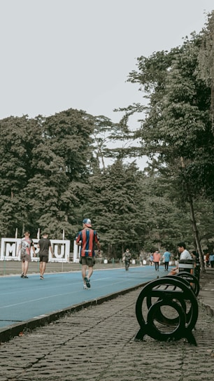 A vibrant outdoor scene featuring a running track surrounded by lush green trees. People are actively walking or jogging on the blue track, some dressed in sportswear. Benches and a decorative sculpture are visible alongside the track. The setting appears lively and recreational.