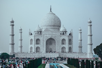 A grand white marble mausoleum with a large dome and four tall minarets, set against a pale sky. The foreground features a rectangular reflecting pool, neatly lined with dark green trees and colorful flowers. A large congregation of people, both locals and tourists, is gathered across the area, highlighting its popularity.