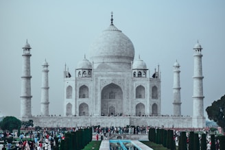 A grand white marble mausoleum with a large dome and four tall minarets, set against a pale sky. The foreground features a rectangular reflecting pool, neatly lined with dark green trees and colorful flowers. A large congregation of people, both locals and tourists, is gathered across the area, highlighting its popularity.