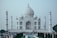 A grand white marble mausoleum with a large dome and four tall minarets, set against a pale sky. The foreground features a rectangular reflecting pool, neatly lined with dark green trees and colorful flowers. A large congregation of people, both locals and tourists, is gathered across the area, highlighting its popularity.