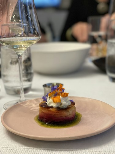 A gourmet dish is elegantly plated on a pale pink plate with layers of thinly sliced vegetables topped with a dollop of cream and edible flowers. A glass of white wine is partially visible in the foreground, suggesting a dining setting. A white bowl and blurred figures are in the background, contributing to a fine dining atmosphere.