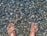 A pair of feet submerged in clear water with a bed of smooth, multicolored pebbles visible underneath. The water creates a shimmering effect on the skin and stones.