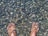A pair of feet submerged in clear water with a bed of smooth, multicolored pebbles visible underneath. The water creates a shimmering effect on the skin and stones.