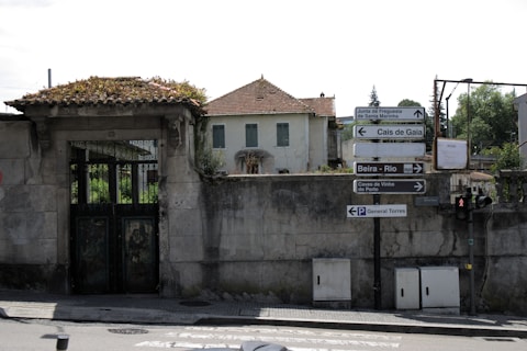 A rundown building with a tiled roof and worn exterior walls, surrounded by an old stone wall. Various street signs point in multiple directions, indicating locations like Cais de Gaia and Beira-Rio. A traffic light and pedestrian crossing are present.