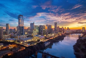 A lively cityscape at sunset with iconic architecture and glowing lights reflecting on the river.