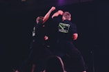 Two individuals are performing martial arts on a dimly lit stage. They are wearing black uniforms with neon green logos on the back labeled 'Team Infinity'. One person is throwing a punch while the other is sidestepping or responding to the movement. The focus is on dynamic action and physical combat training.