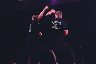 Two individuals are performing martial arts on a dimly lit stage. They are wearing black uniforms with neon green logos on the back labeled 'Team Infinity'. One person is throwing a punch while the other is sidestepping or responding to the movement. The focus is on dynamic action and physical combat training.